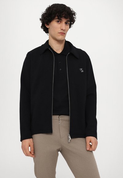 Svart zip-up jacka med krage, som har en svag lyster. Bärs över en svart polo och matchas med beige byxor. Diskret logotypdetalj.