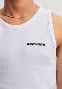 Personne portant un débardeur blanc côtelé avec le texte « "PEGADOR" » et un collier en chaîne argentée, visible de l’épaule au haut de la poitrine.