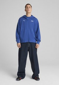 Plava zip majica s kengurskim džepovima, nošena preko plave košulje, u kombinaciji s labavim tamnim trapericama i crnim tenisicama.