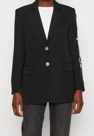 Zwart op maat gemaakte blazer met twee zilveren knopen, klepzakken en witte tekst op de rechtermouw, gedragen over een witte top en donkere spijkerbroek.