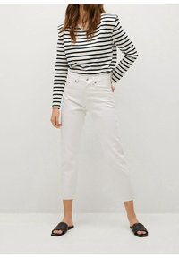 Jeans blancos cortos combinados con una camiseta de rayas en blanco y negro de manga larga. La modelo lleva sandalias negras tipo slide. Fondo plano.