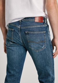 Vaqueros de mezclilla de un azul oscuro con corte ajustado, que cuentan con varios bolsillos, costuras claras y una etiqueta de cuero marrón de la marca en la cintura.