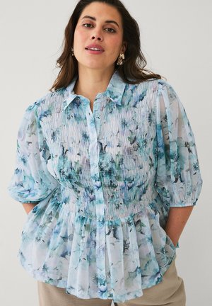 Kvinde iført en blå og grøn blomstret smockbluse med puffede trekvartærmer og beige bukser mod en ensfarvet baggrund.