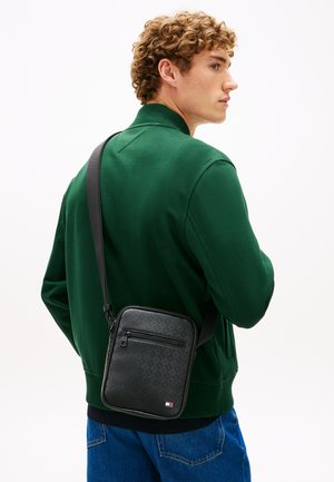 Joven con cabello rizado que lleva una chaqueta verde y un bolso negro texturizado de bandolera con bolsillo frontal con cremallera.