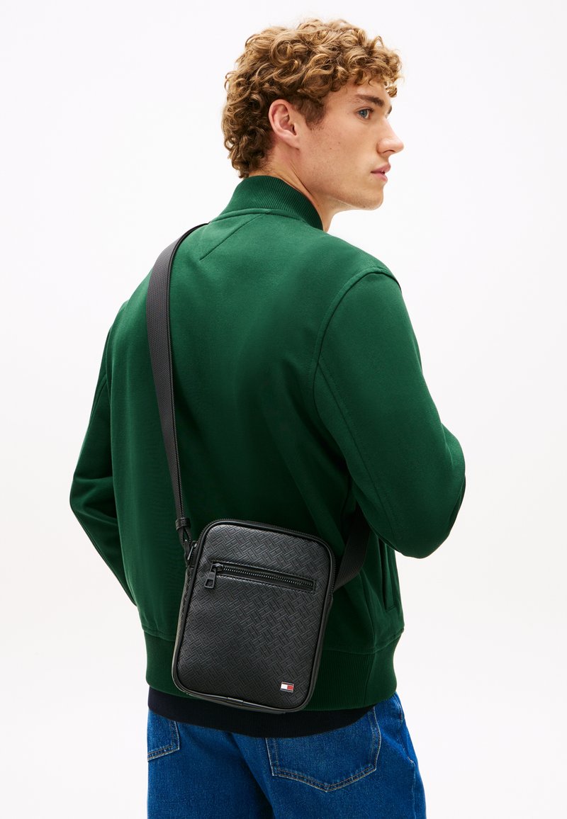 Νέος άνδρας με κατσαρά μαλλιά, φορώντας πράσινο μπουφάν και μαύρη τσάντα χιαστί με υφή και τσέπη με φερμουάρ μπροστά.