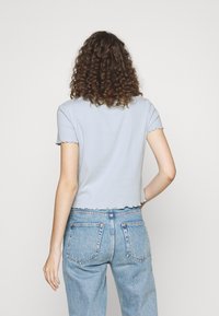 Žena s kudrnatými vlasy, oblečená v lehkém modrém tričku s krátkým rukávem a džínech s vysokým pasem v světle modré barvě, otočená zády k fotoaparátu.