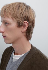 Vista profilo di una persona con capelli biondo chiaro che indossa un maglione grigio a scollo a v sovrapposto a un cardigan verde oliva scuro, con pelle liscia e trame sottili.