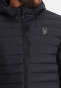 Zwarte pufferjack met een gekleed textuur, voorzien van een volledige rits en een klein spinnenlogo op de bovenste linkerzijde.