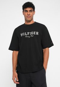 Čierne tričko s krátkym rukávom s textúrovanou tkaninou, okrúhlym výstrihom a potlačou loga "HILFIGER Racing Club" v bielej farbe na prednej strane.