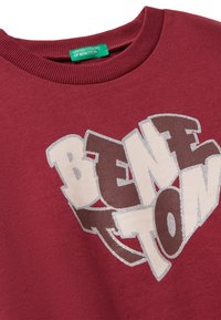 Sudadera color marrón con cuello de canalé y diseño de texto en forma de corazón "Benetton" en letras beige y marrones con contorno de purpurina plateada.