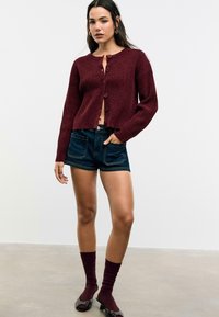 Bordo pleteni kardigan s okruglim izrezom i detaljima od dugmadi, uparen s tamnim traper kratkim hlačama. Model nosi usklađene bordo čarape i cipele.