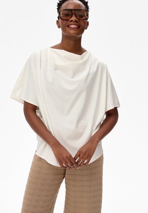 Uśmiechnięta osoba w dużych brązowych okularach przeciwsłonecznych, białym, luźnym topie z krótkim rękawem oraz beżowych, teksturowanych dzianinowych spodniach na białym tle.