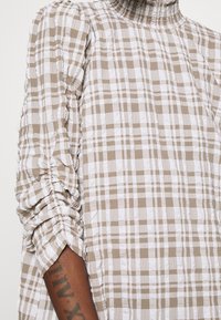 Checkered beige and white blouse with a high neckline, gathered sleeves, and textured fabric. Wrinkled finish adds dimension and interest.