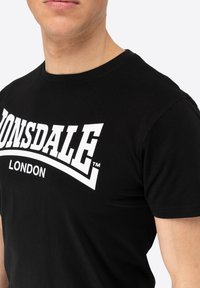Melna kokvilnas T-krekls ar drosmīgu balto "LONSDALE LONDON" logotipu pie krūtīm. Klasisks apaļais kakla dizains, īsas piedurknes.