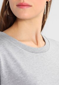 Jasnoszara bluza z ribbanym dekoltem, o miękkiej fakturze. Duże geometryczne kolczyki w kształcie kółek z złotego metalu podkreślają całość.
