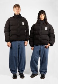 Black puffer jackets with zip pockets and a logo on the chest, paired with loose blue denim jeans and black footwear.