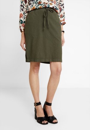 Kobieta ubrana w oliwkową spódnicę do kolan z troczkiem w talii, zestawioną z czarnymi sandałami na obcasie i kwiecistą, długorękawową bluzką.