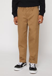 Tan kleurige katoenen broek met een rechte snit, voorzien van een voorzak en een ritssluiting. Gepaard met zwarte sneakers met witte veters en accenten.