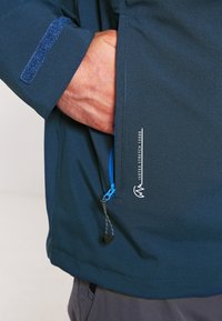Veste imperméable bleue avec une poche zippée dotée d'une fermeture éclair bleue vif, des détails texturés sur les manches et un logo de marque visible.