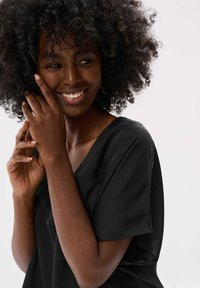 Zwarte blouse met korte mouwen en een V-hals ontwerp, met een gladde textuur en subtiele glans, gedragen door een model met krullend haar in een natuurlijke pose.