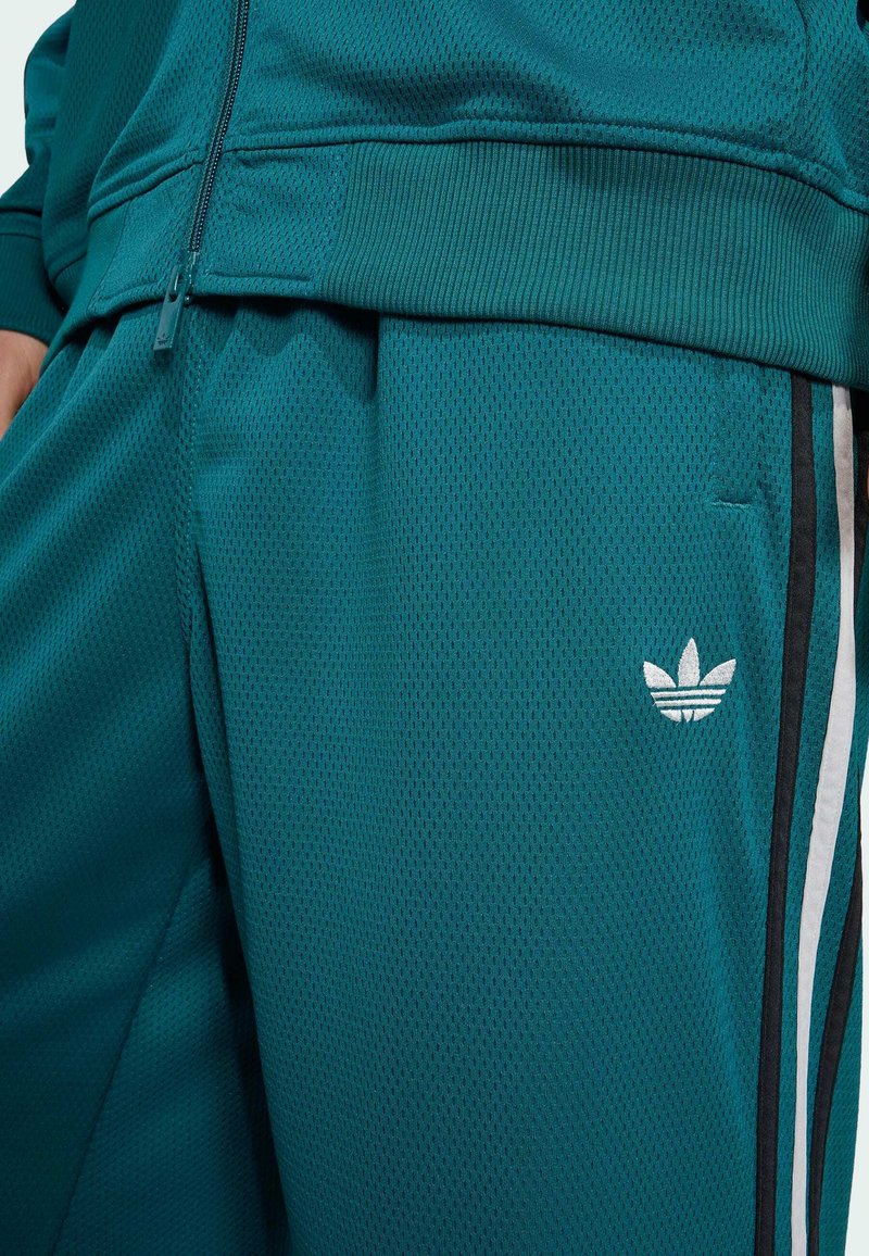 Türkisfarbene Sporthose und Jacke mit schwarz-weißen Seitenstreifen, mit weißem Dreiblatt-Logo in der Nähe der Tasche.