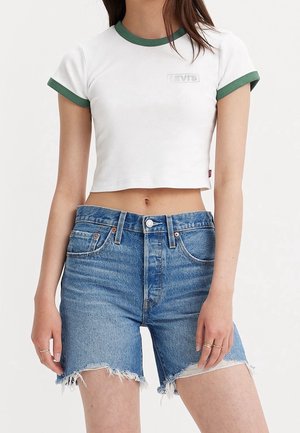 Kobieta ubrana w biały krótki t-shirt z zielonym wykończeniem i wysokowysokościowe niebieskie dżinsowe szorty z postrzępionymi brzegami.