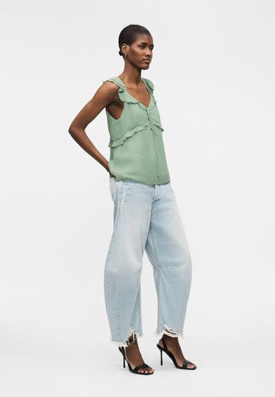 Γυναίκα που στέκεται με τα χέρια στις πίσω τσέπες, φοράει πράσινο αμάνικο τοπ με βολάν, φαρδιά ανοιχτά μπλε τζιν με φθορές και μαύρα σανδάλια με ψηλό τακούνι.