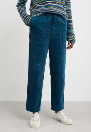 Personne portant un pantalon en velours côtelé bleu sarcelle avec des poches avant, un pull rayé multicolore et des baskets blanches, debout sur un sol clair.