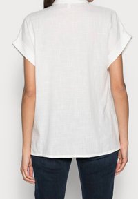 Biała bluzka z krótkimi rękawami o fakturowanym wzorze, luźny krój i zaokrąglony dół. Materiał wydaje się lekki i casualowy. Połączona z ciemnymi dżinsami.