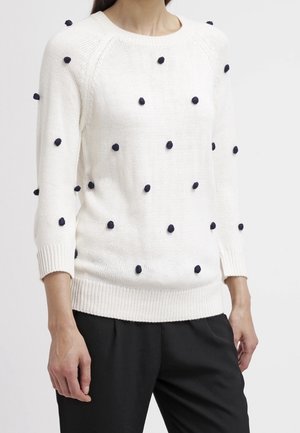 Person trägt einen weißen Strickpullover mit schwarzen, strukturierten Punktakzenten und eine schwarze Hose, stehend vor einem schlichten, hellen Hintergrund.