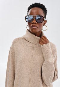 Beżowy sweter dzianinowy z wysokim kołnierzem i prążkowanymi mankietami. Modelka nosi dużą oprawkę w kolorze żółwiej skorupy oraz złote kolczyki w kształcie kół.