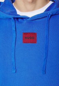 Sudadera con capucha de algodón azul brillante con un parche rectangular rojo etiquetado "HUGO", con cordones y puños de canalé.