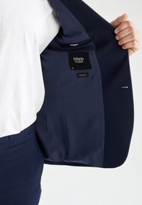 Inuti den marinblå kavajen syns ett slätt tygfoder med en etikett som visar "DeFacto STUDIO, storlek 50, SLIM FIT." Den har en ficka och skräddarsydda sömmar.