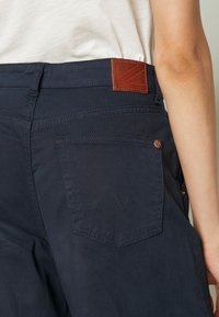Personne portant un pantalon bleu foncé avec une poche arrière et un patch de marque en cuir marron à la taille, montrée de la taille vers le bas.