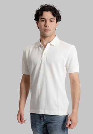 Polo bianco in cotone, con colletto classico, maniche corte e un logo discreto sul petto. Indossato con jeans blu.