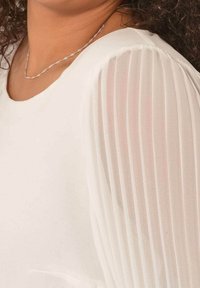 White blouse with softly pleated sheer sleeves, featuring a rounded neckline and smooth texture. Accentuated by a delicate silver necklace.