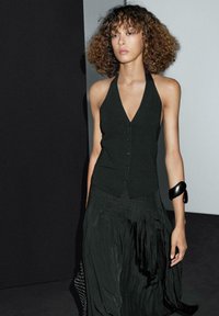 Black halter top with a V-neck and button detail, paired with a flowy black pleated skirt. Worn with a black bangle on the wrist.