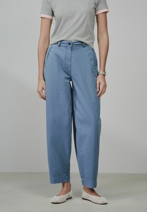 Person iført en lysegrå T-shirt, højtaljede løse blå bukser, hvide nette flade sko og et armbåndsur, stående mod en ensfarvet baggrund.