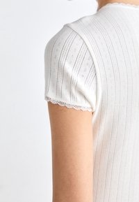 Biała bluzka z krótkim rękawem, z teksturowanym wzorem w pionowe paski i detalami z oczkami, z delikatnym koronkowym wykończeniem u rękawów.