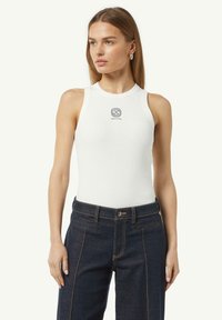 Witte geribbelde tanktop met een hoge halslijn en een mouwloos ontwerp, voorzien van een klein zwart logo op de borst. Gecombineerd met donkere spijkerbroeken.