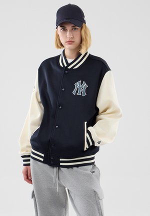 Person iført navy- og cremefarvet varsity-jakke med New York Yankees-logo, navy cap og grå joggingbukser med sidelommer, stående mod en ensfarvet baggrund.