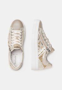 Zapatillas beige con acentos metálicos brillantes, tela texturizada y un panel lateral brilloso. Presenta cordones y una suela de goma.