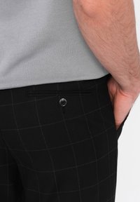 Pantalons à carreaux noirs avec une texture lisse, dotés d'une poche arrière avec un détail de bouton, portés avec une chemise colée gris clair.