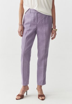 Vrouw draagt op maat gemaakte lichtpaarse broek, wit topje, bruine sandalen met hoge hak, gouden armband en een dunne polstattoo zichtbaar.