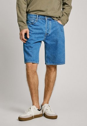 Pantalones cortos de mezclilla en un tono azul claro, con un corte clásico, trabillas para cinturón y bolsillos delanteros y traseros, combinados con zapatillas blancas y grises.