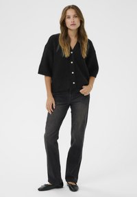 Black knitted cardigan with short sleeves, featuring large buttons and ribbed hem, paired with dark jeans and black flats, against a white background.