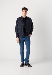 Chaqueta marina con dos bolsillos frontales, llevada sobre una camiseta polo negra, combinada con vaqueros azules y zapatillas negras. Tejido suave con un corte relajado.