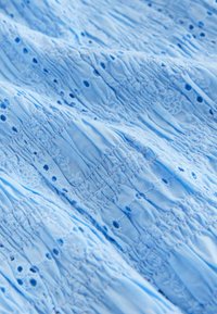 Šviesiai mėlyna audinys su raižytu siuvinėjimu ir raukiniais, su mažais išpjovų elementais ir gėlių raštais visame paviršiuje.