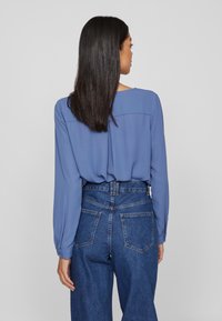 Blusa azzurro chiaro a maniche lunghe, in tessuto morbido, con scollatura rotonda e design con yoke sul retro, abbinata a jeans in denim a vita alta.