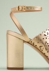 Gold, heeled sandal with a wide block heel, cut-out floral design on the upper, and adjustable strap with a metallic buckle.